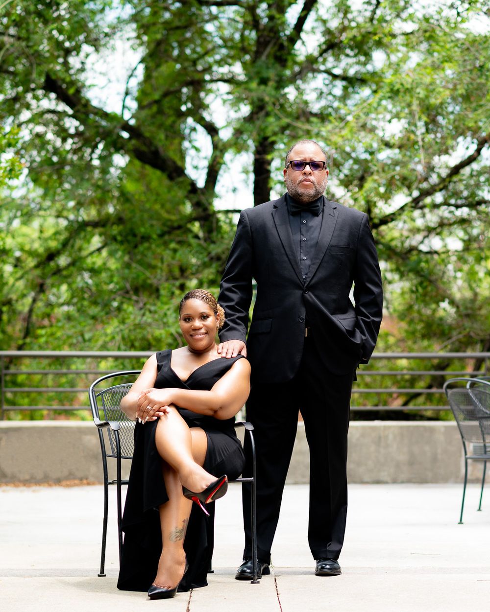 Couple in formal black attire poses with outdoor furniture.