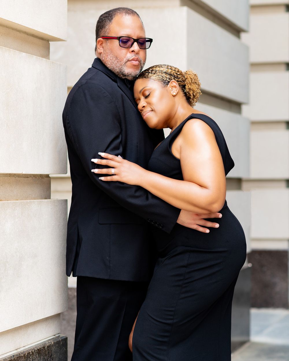 Couple in black formal wear embrace against light stone wall.