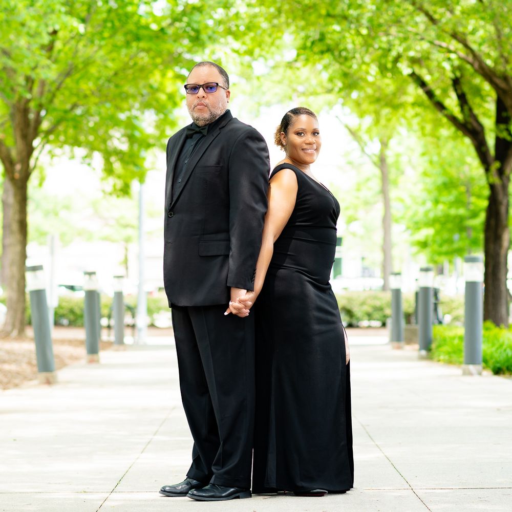 Couple in matching black formal attire poses on tree-lined walkway.