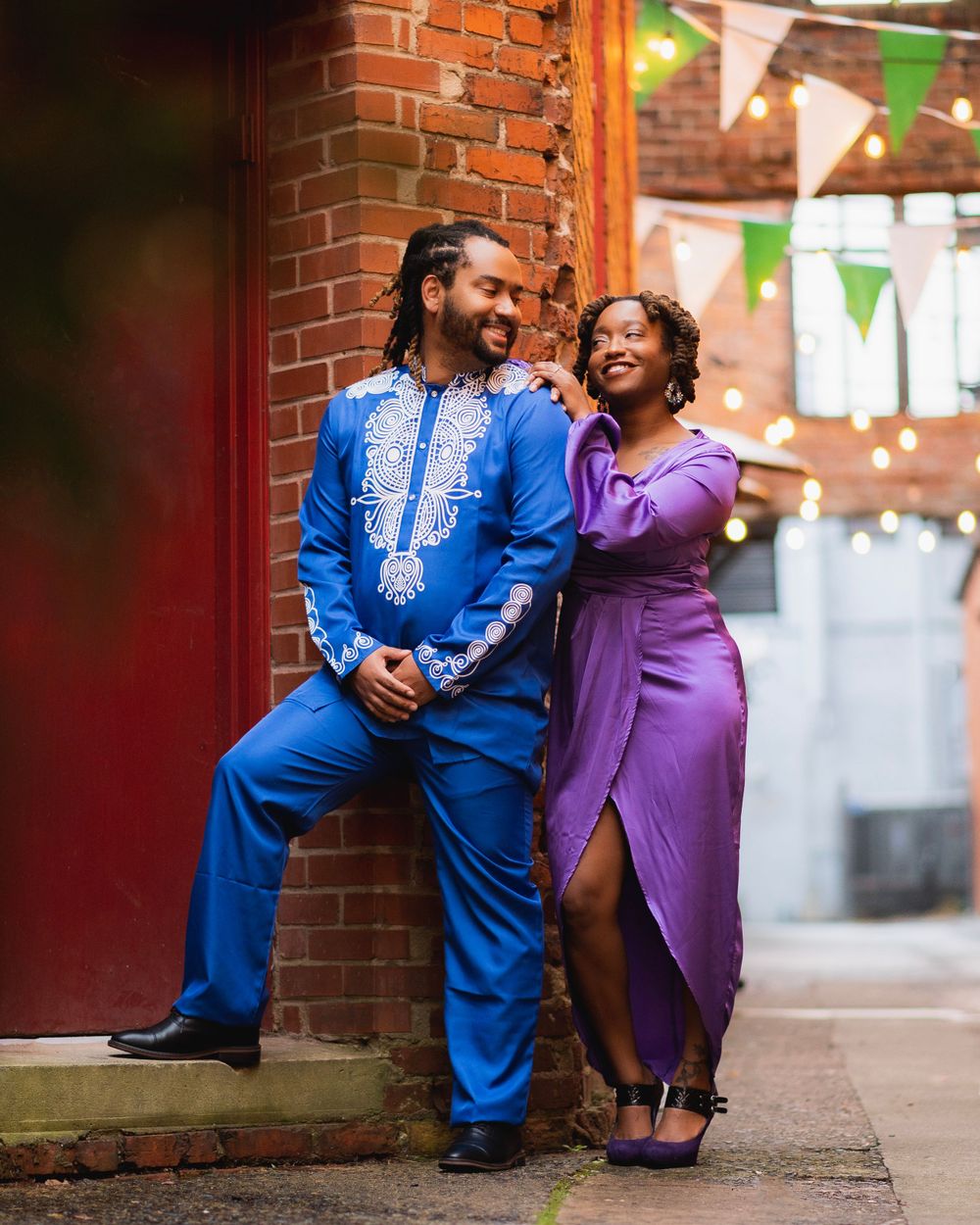 Couple in elegant purple dress and blue traditional outfit lean against brick wall under festive lighting.