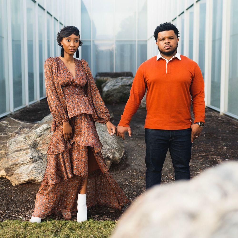 Couple poses between shipping containers, wearing orange sweater and brown tiered dress.