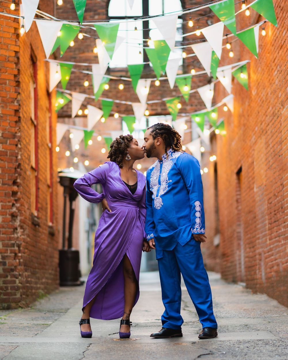 Couple shares romantic moment under green and white bunting flags in brick alleyway.
