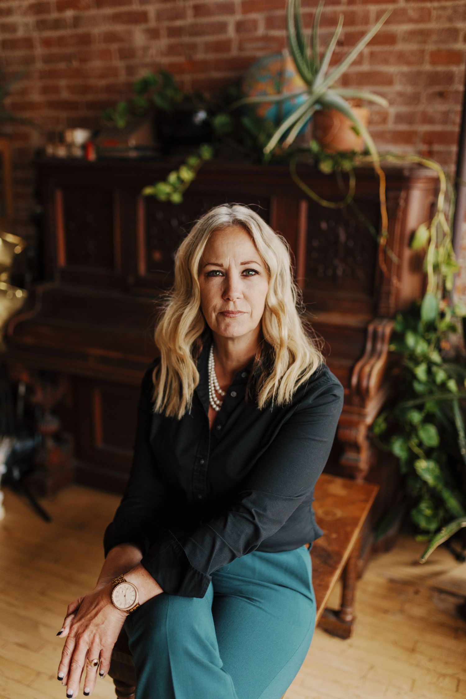 A professional portrait in front of brick wall with indoor plants and vintage furniture.