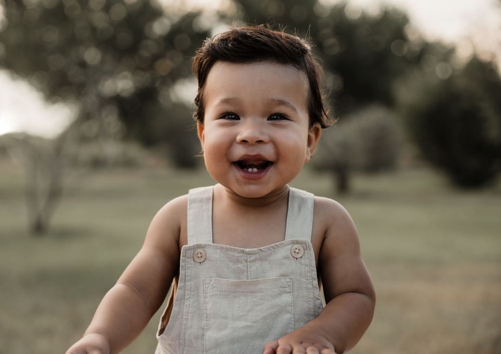 Happy toddler in white overalls smiles brightly in outdoor garden setting.