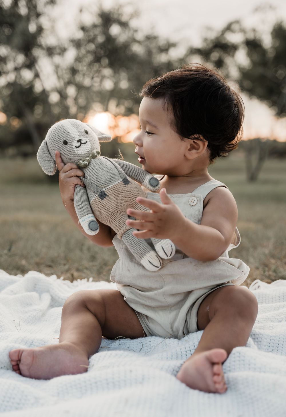 Baby in light overalls plays with gray stuffed bunny toy while sitting on white blanket outdoors.