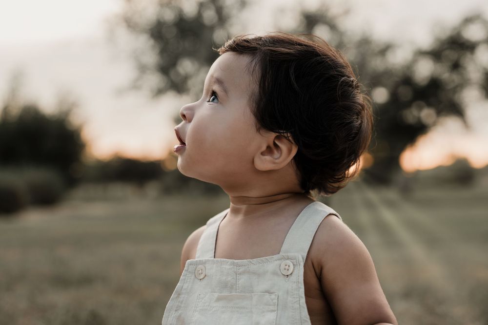 Baby in light colored overalls gazes upward during golden hour outdoor portrait session.