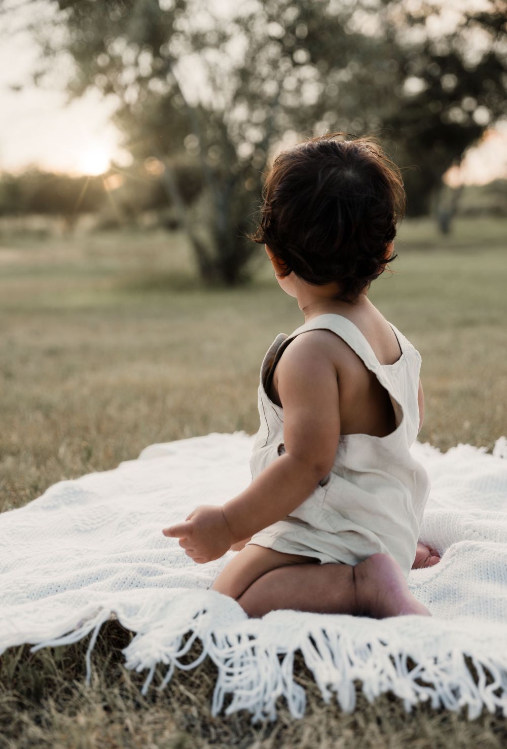 Baby in white tank top sits on textured blanket during sunset in grassy field.