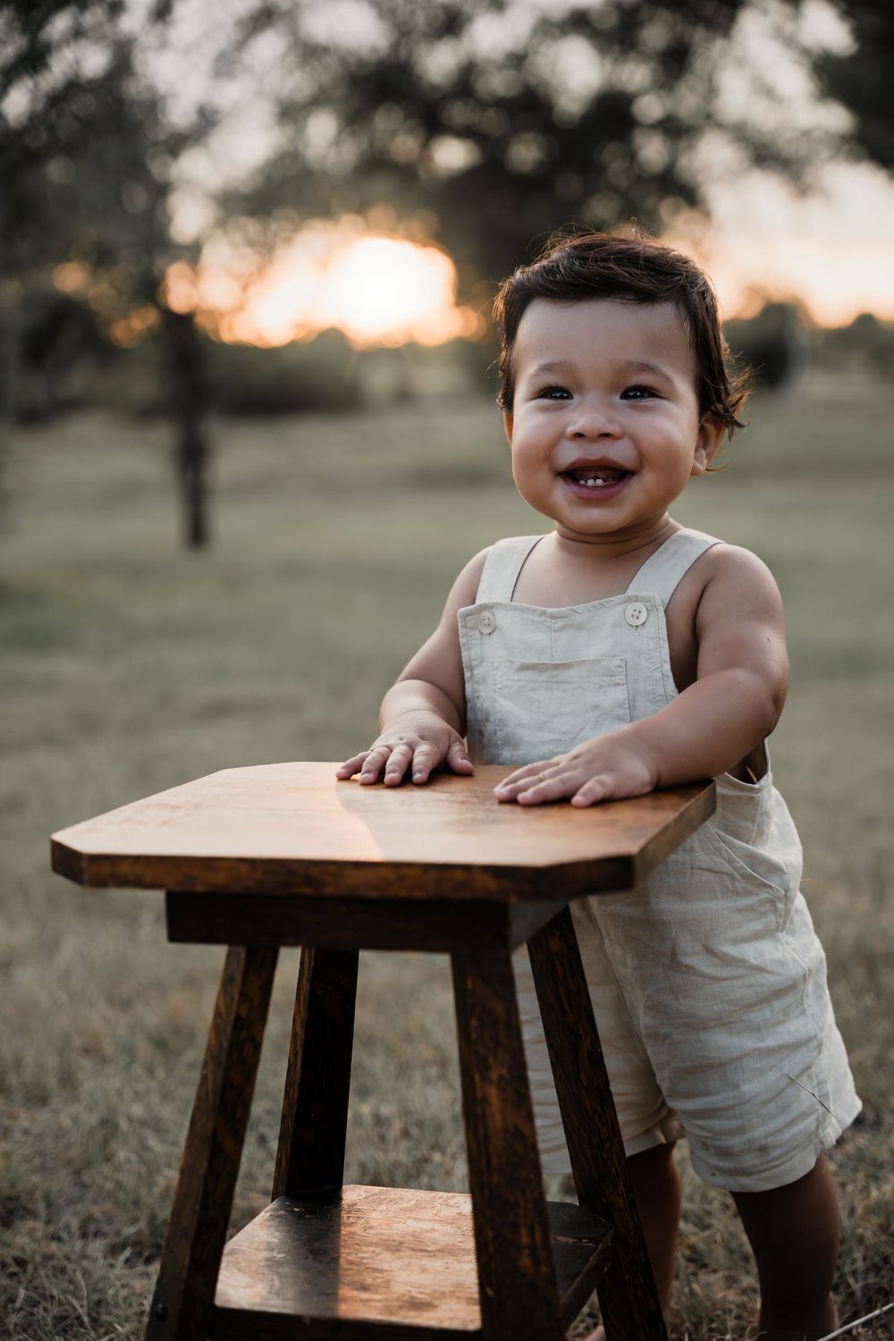 Laughing baby in overalls stands at wooden stool during sunset outdoor photography session.