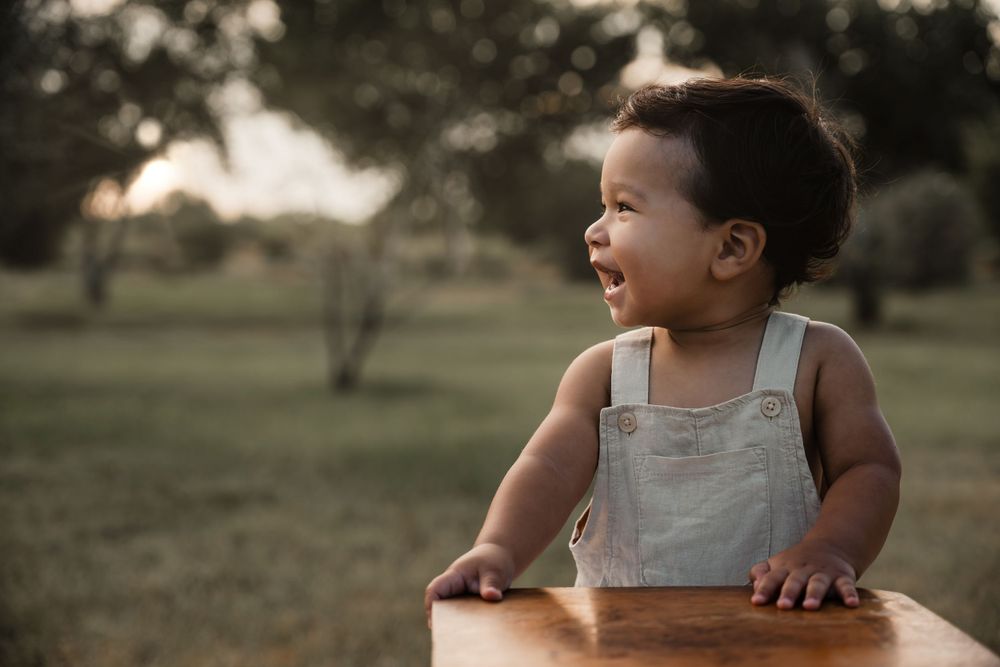 Toddler in light gray overalls smiles while leaning on wooden furniture during golden hour outdoor photoshoot.