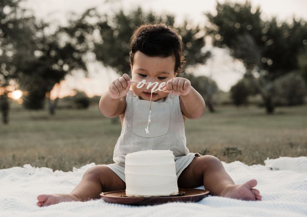 Baby in white overalls plays while sitting on blanket during outdoor sunset session.
