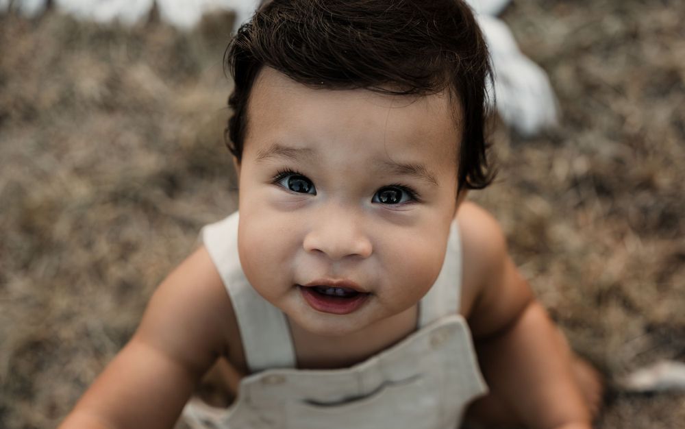 Close-up portrait of smiling baby wearing light gray overalls outdoors.
