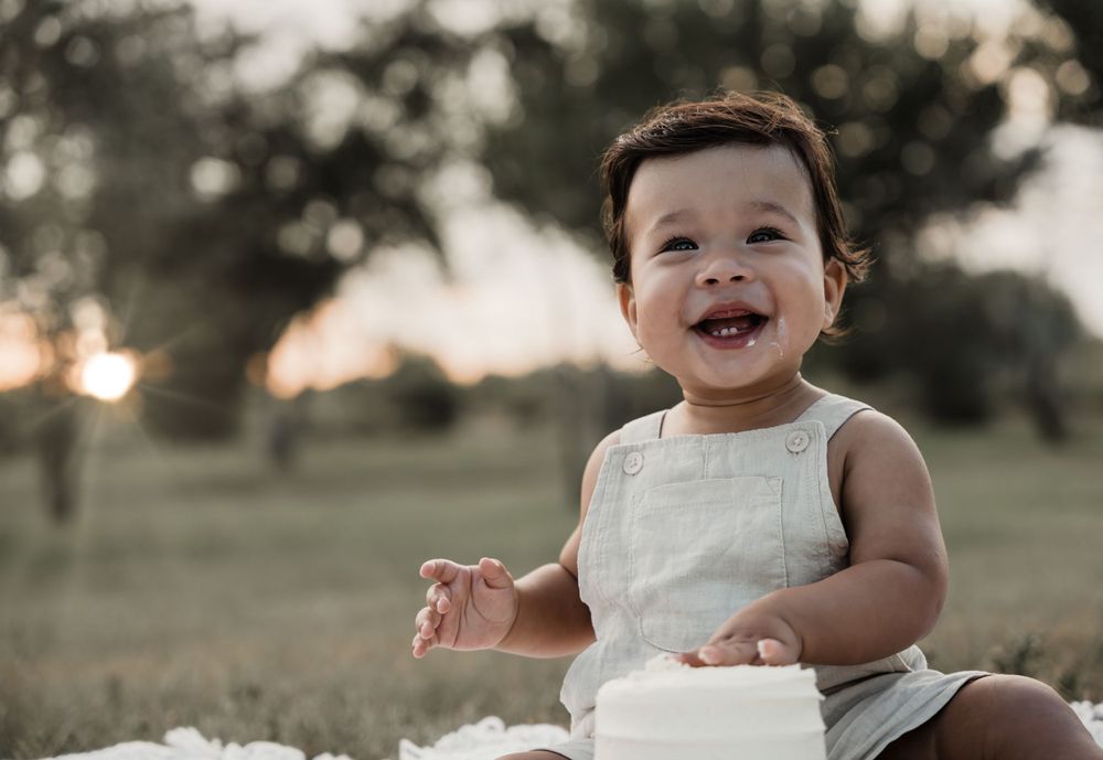 Happy baby in white overalls laughs while sitting on blanket during outdoor sunset session.