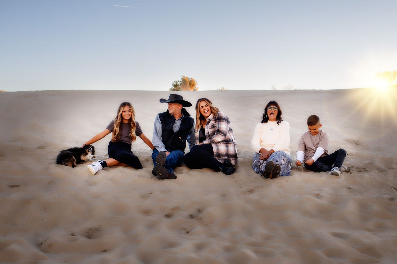 Group sitting together on sand dunes at sunset with a dog.