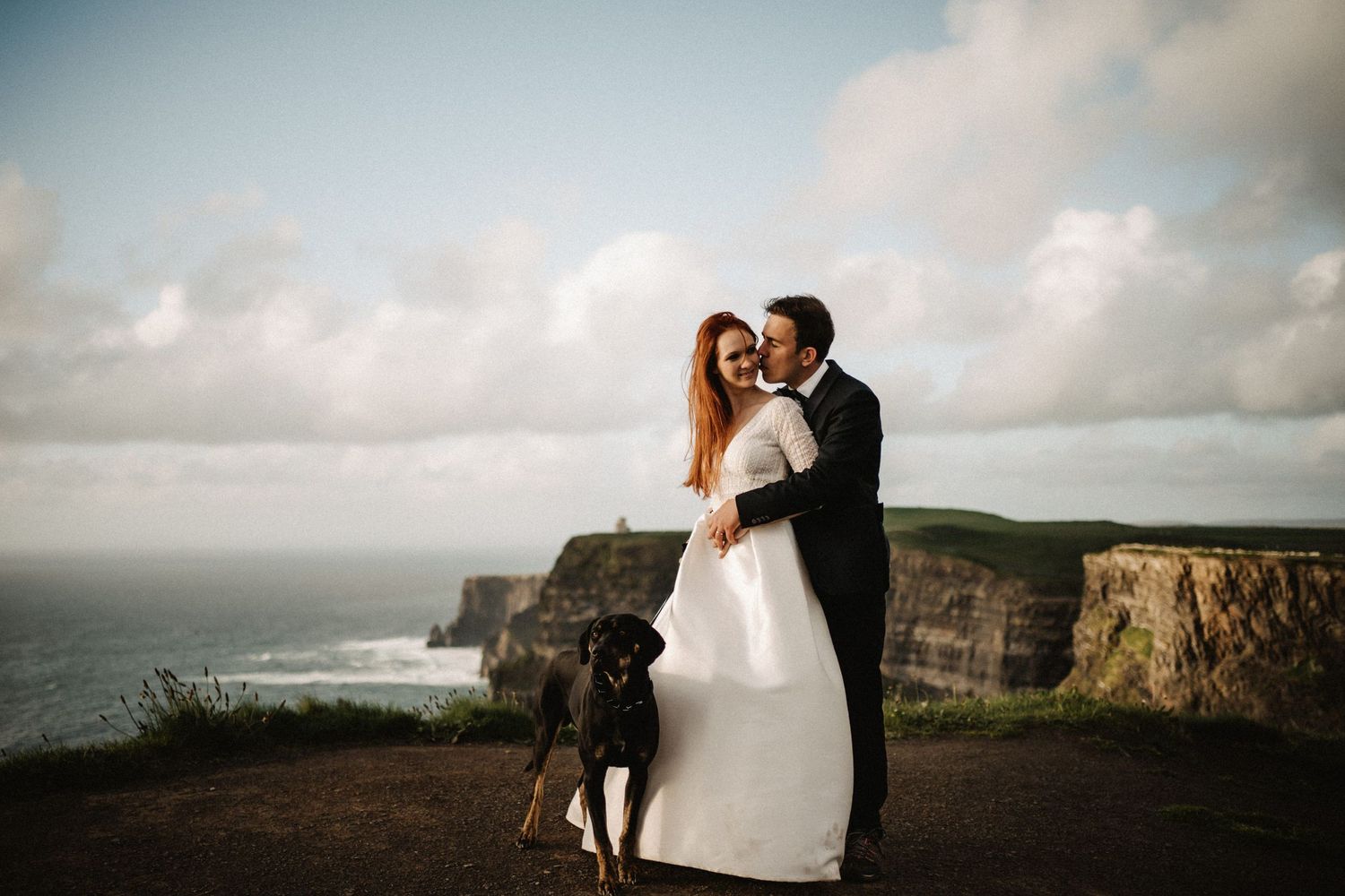 A dramatic wedding portrait at the Cliffs of Moher with a couple and their dog against the ocean view.