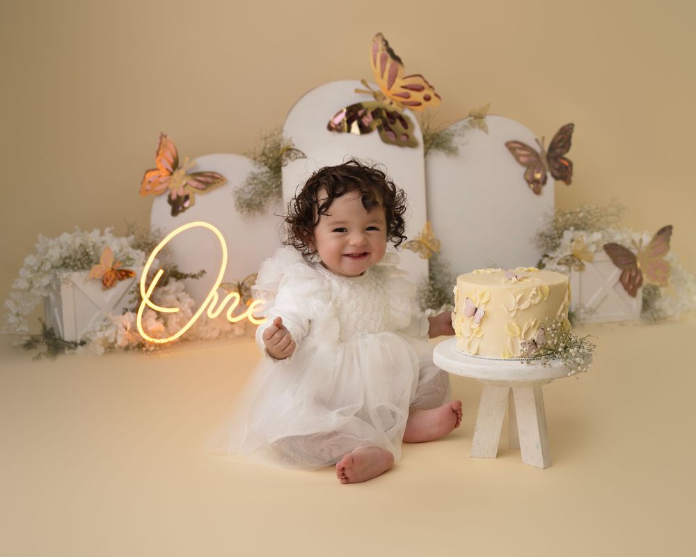 Baby in white dress with butterfly decorations and cake setup for milestone photo.