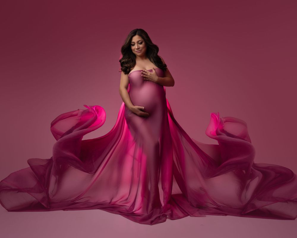 Expectant mother in flowing pink gown against burgundy backdrop with dramatic fabric movement.