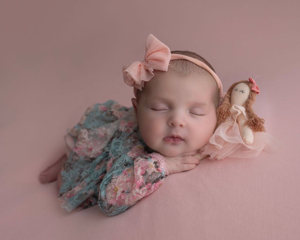 Sleeping baby in floral dress with pink bow headband and small doll on pink background.