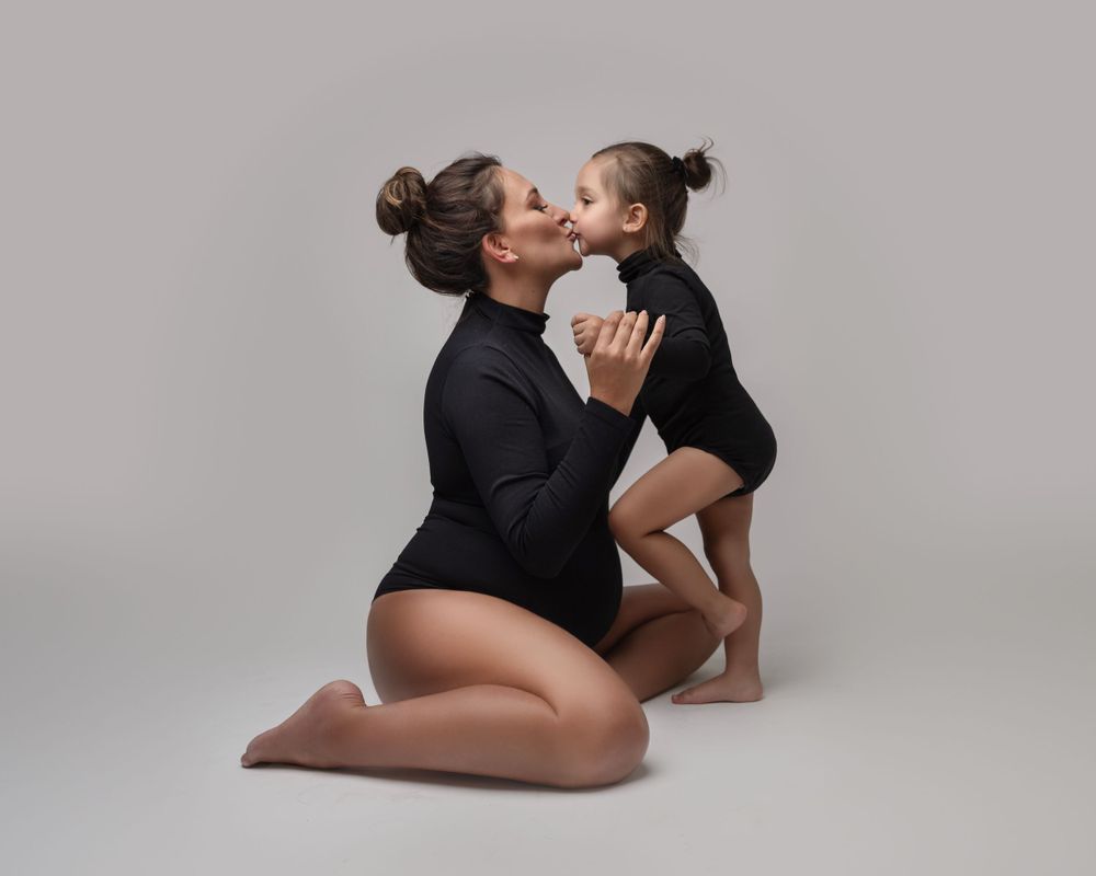 Mother and child in matching black leotards sharing tender moment against grey backdrop.