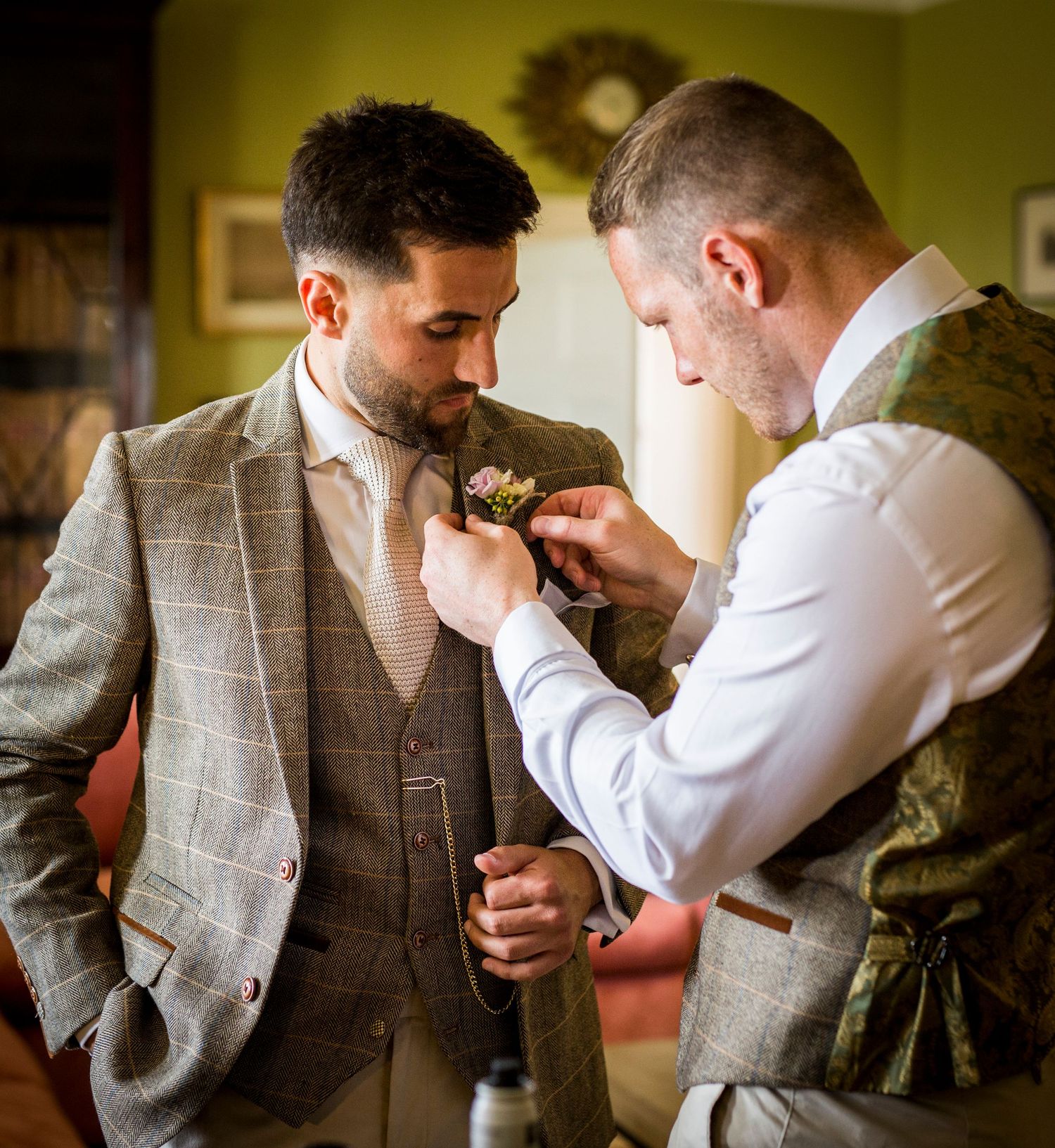 Two people in formal attire help adjust a boutonniere on a gray checkered suit jacket during wedding preparations.
