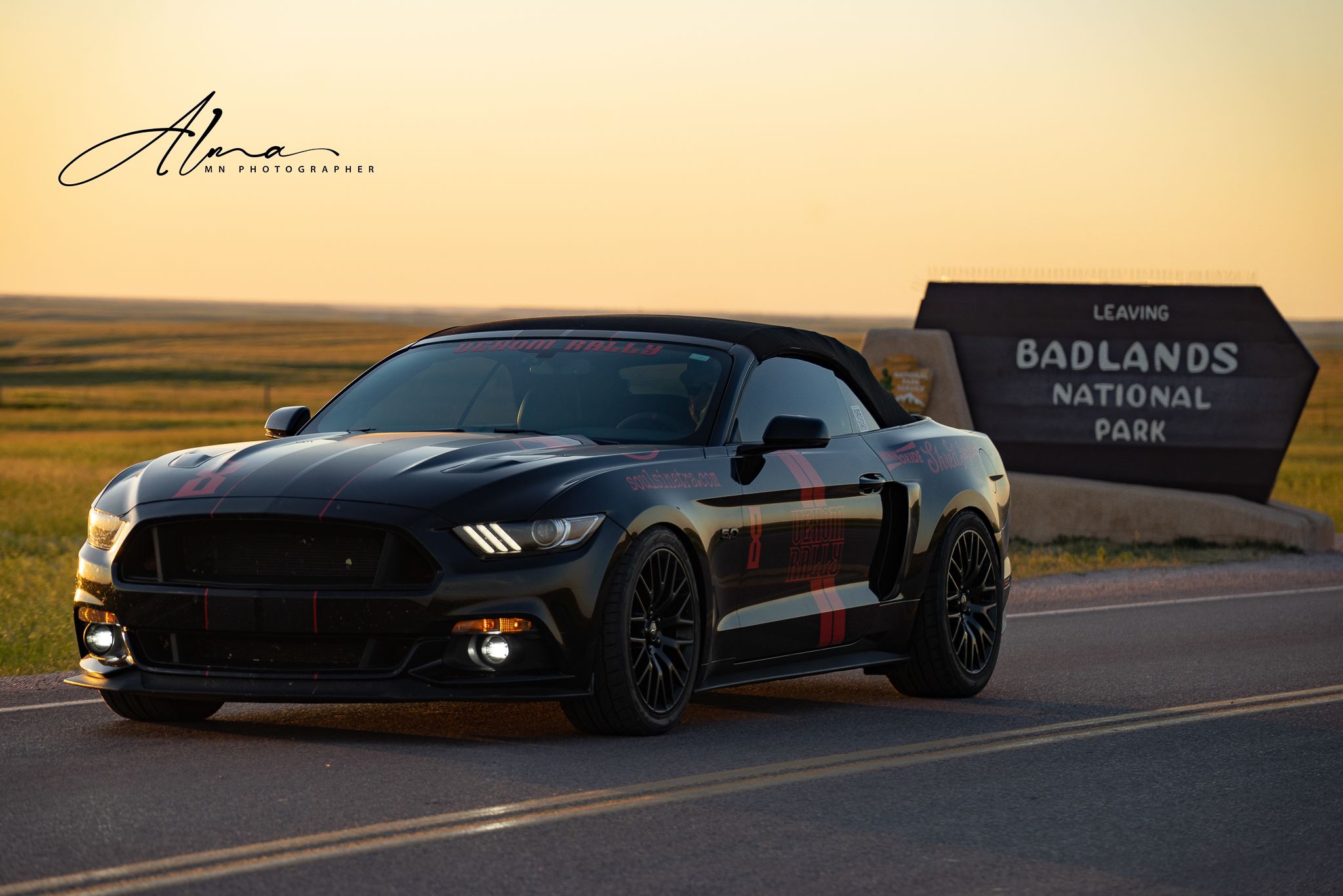 Black Ford Mustang parked at sunset near Badlands National Park entrance sign.