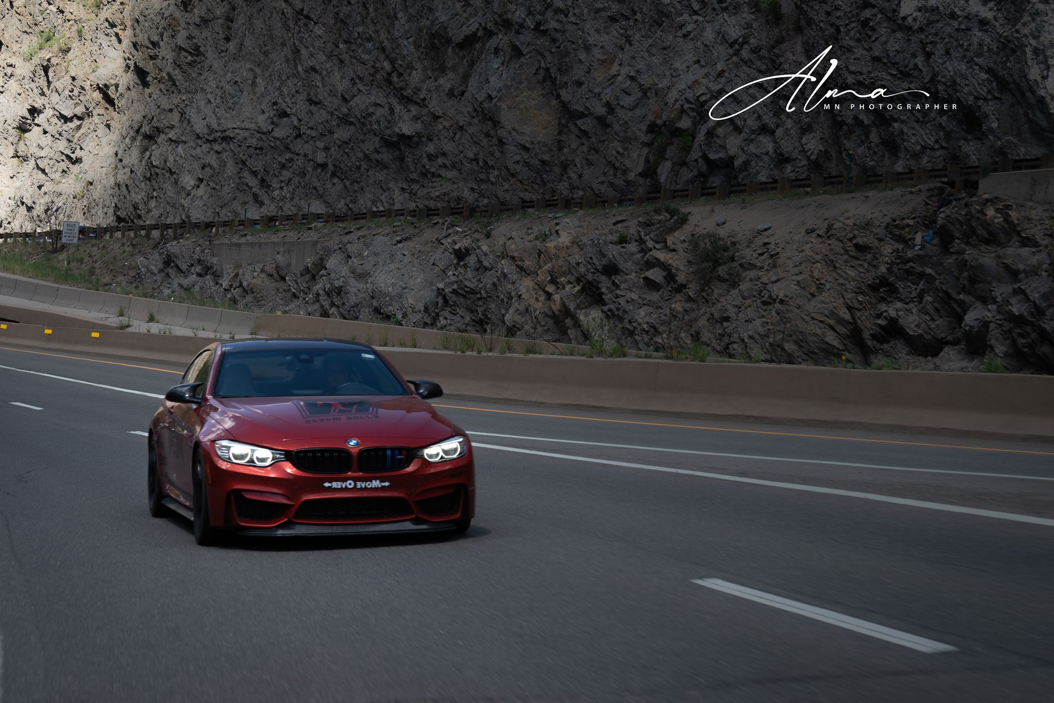 Red sports car driving on mountain road with rocky cliff wall in background.