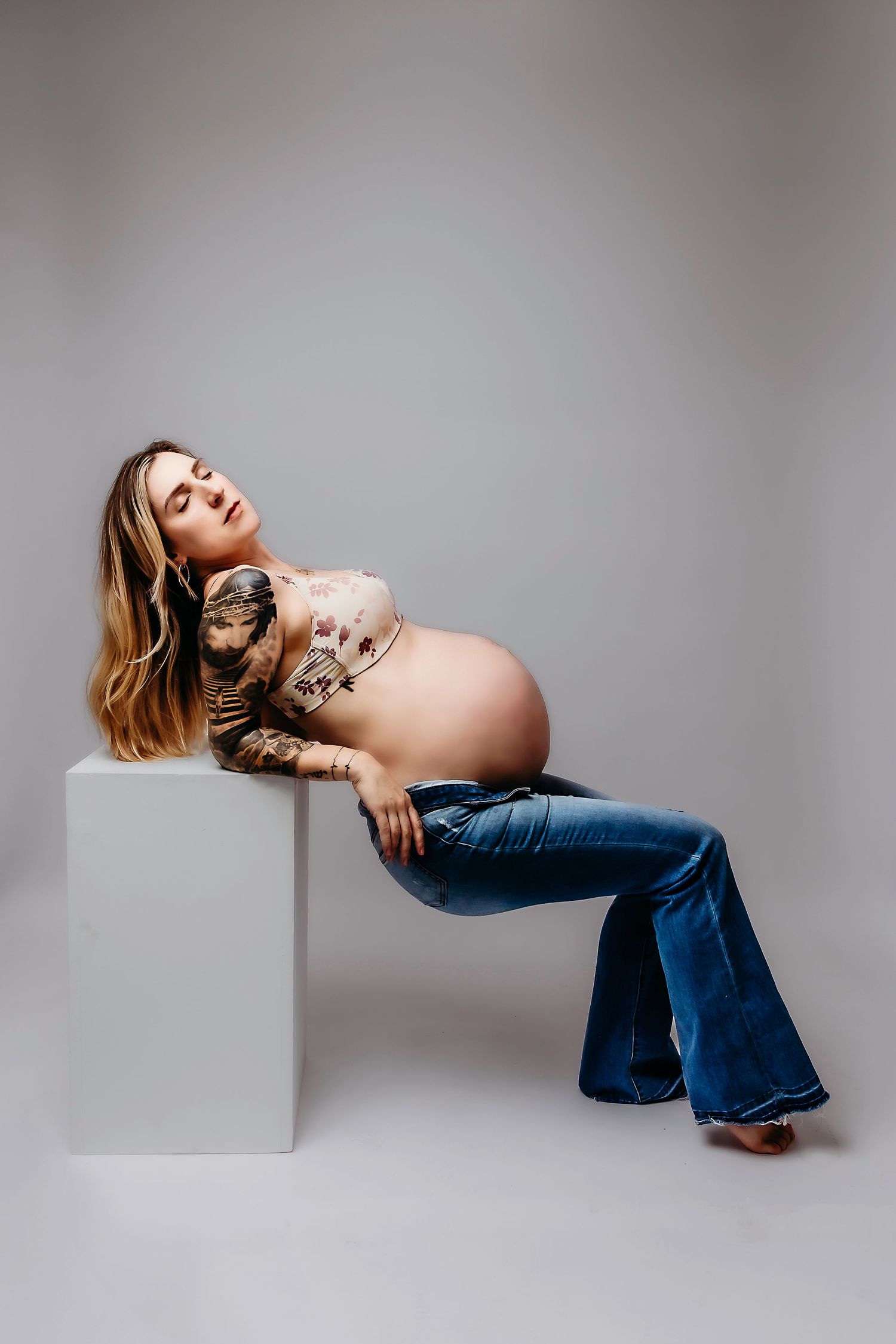 Maternity portrait showing woman in jeans reclining on white cube with visible tattoos.