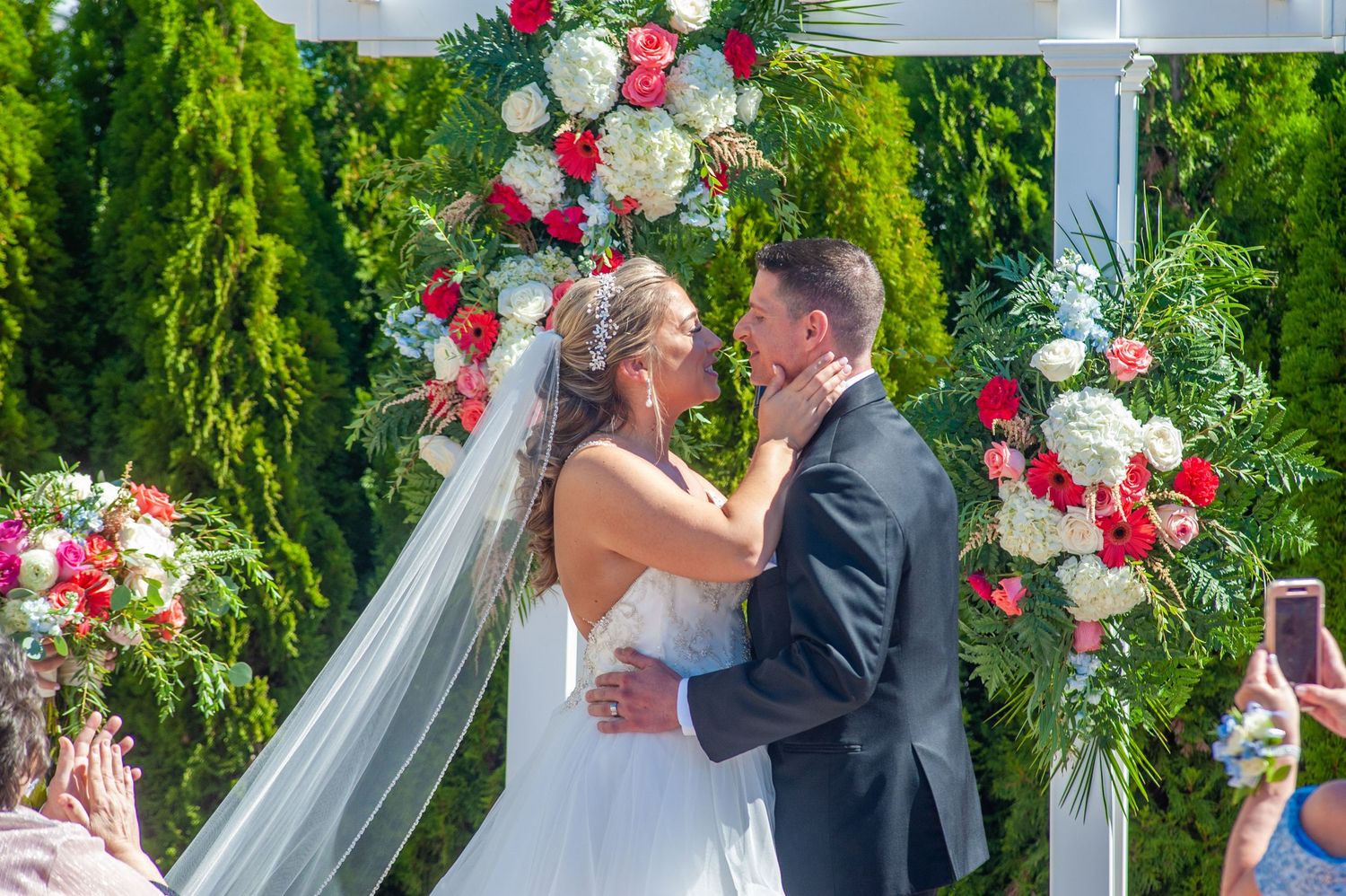 Outdoor wedding ceremony under floral arch with pink and white roses against green garden backdrop.