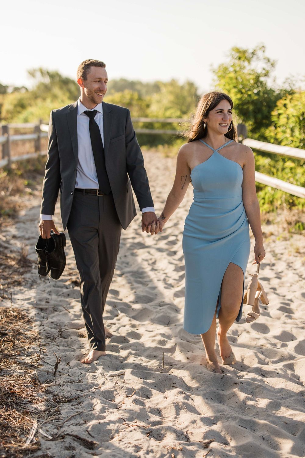 Couple walking barefoot on sandy beach path, dressed elegantly with formal attire.