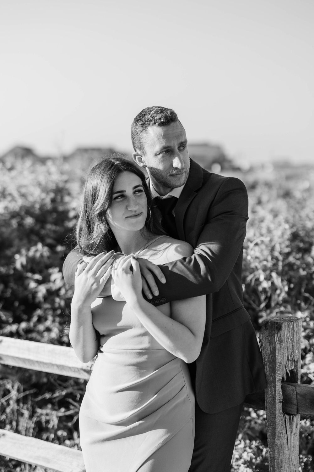 Black and white portrait of couple embracing by rustic fence in natural setting.