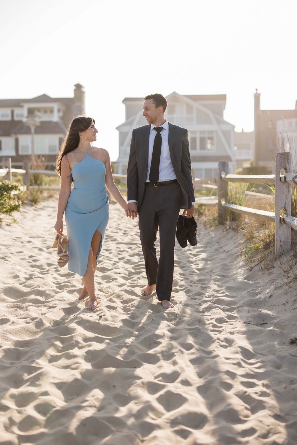 Elegant couple walking barefoot on sandy beach path at sunset near coastal homes.
