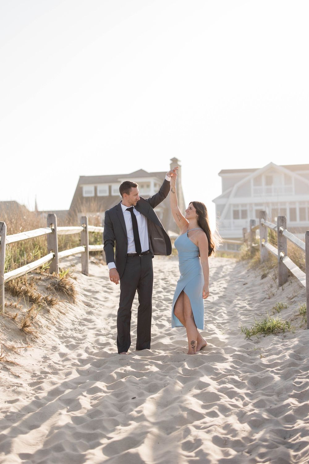 A couple in formal attire share a joyful moment on a sandy beach pathway between coastal homes at sunset.