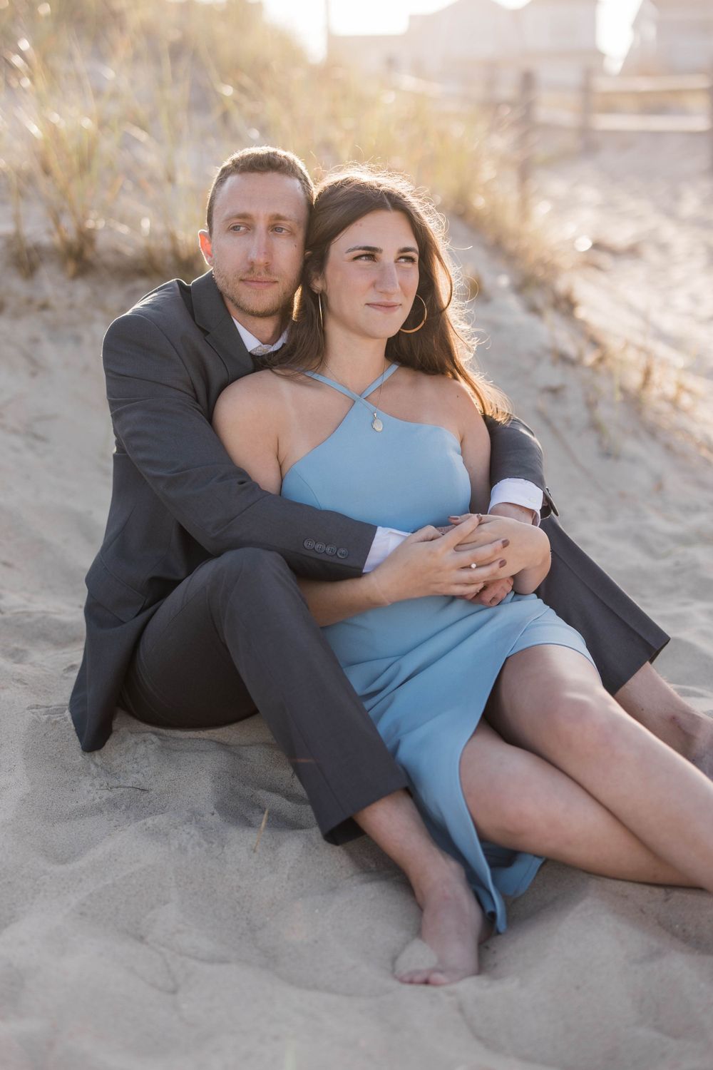 A couple in formal attire sits together on beach sand during golden hour.