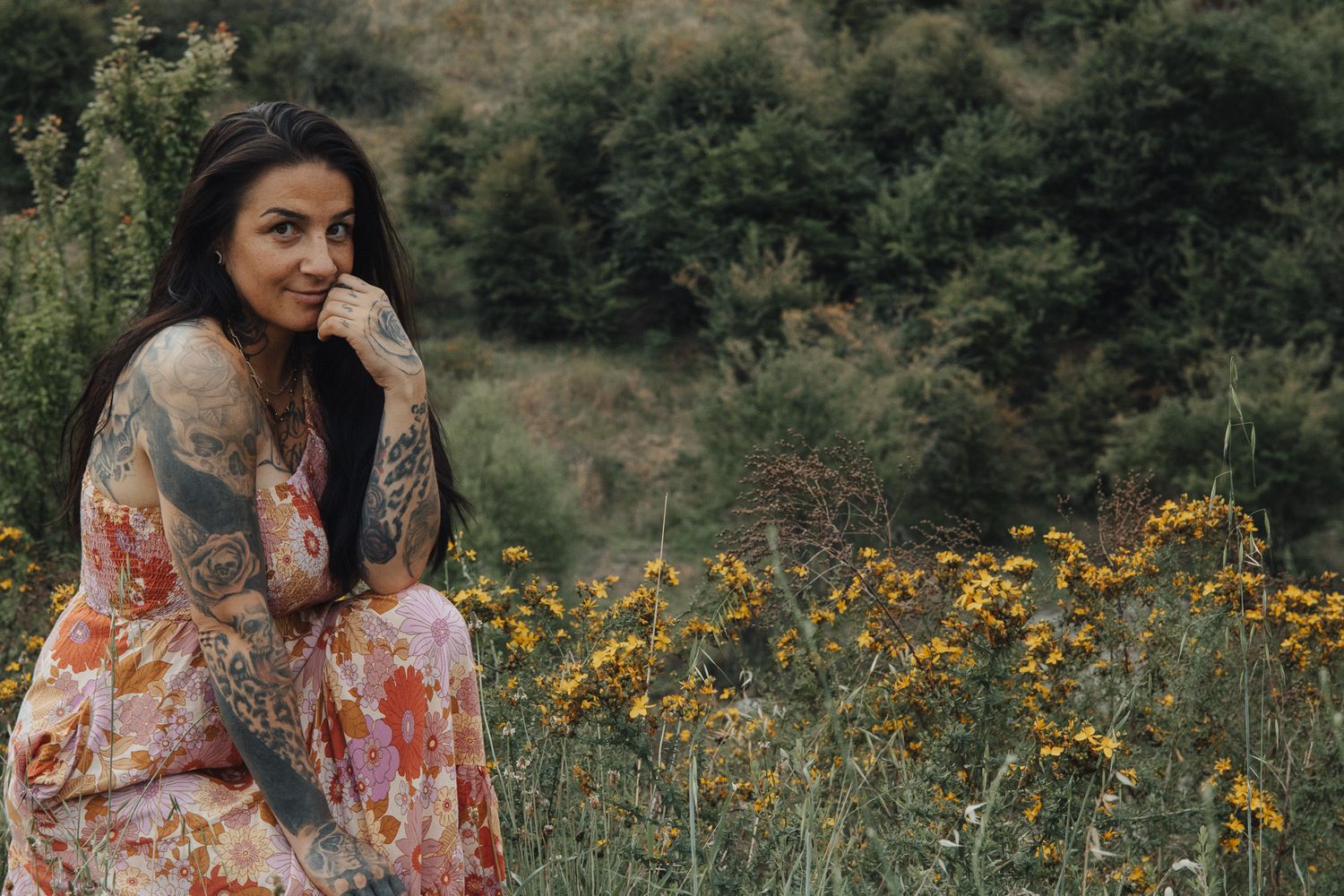 Woman in floral dress sits thoughtfully among yellow wildflowers in natural setting.