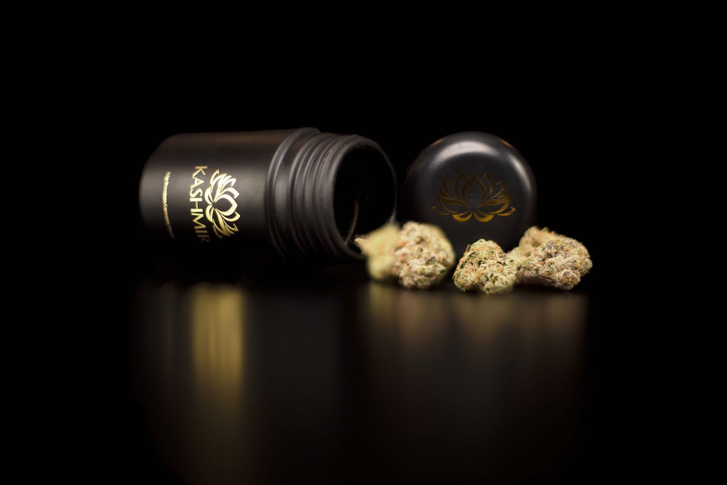 Black cannabis container tipped over with marijuana buds spilling out on reflective surface.