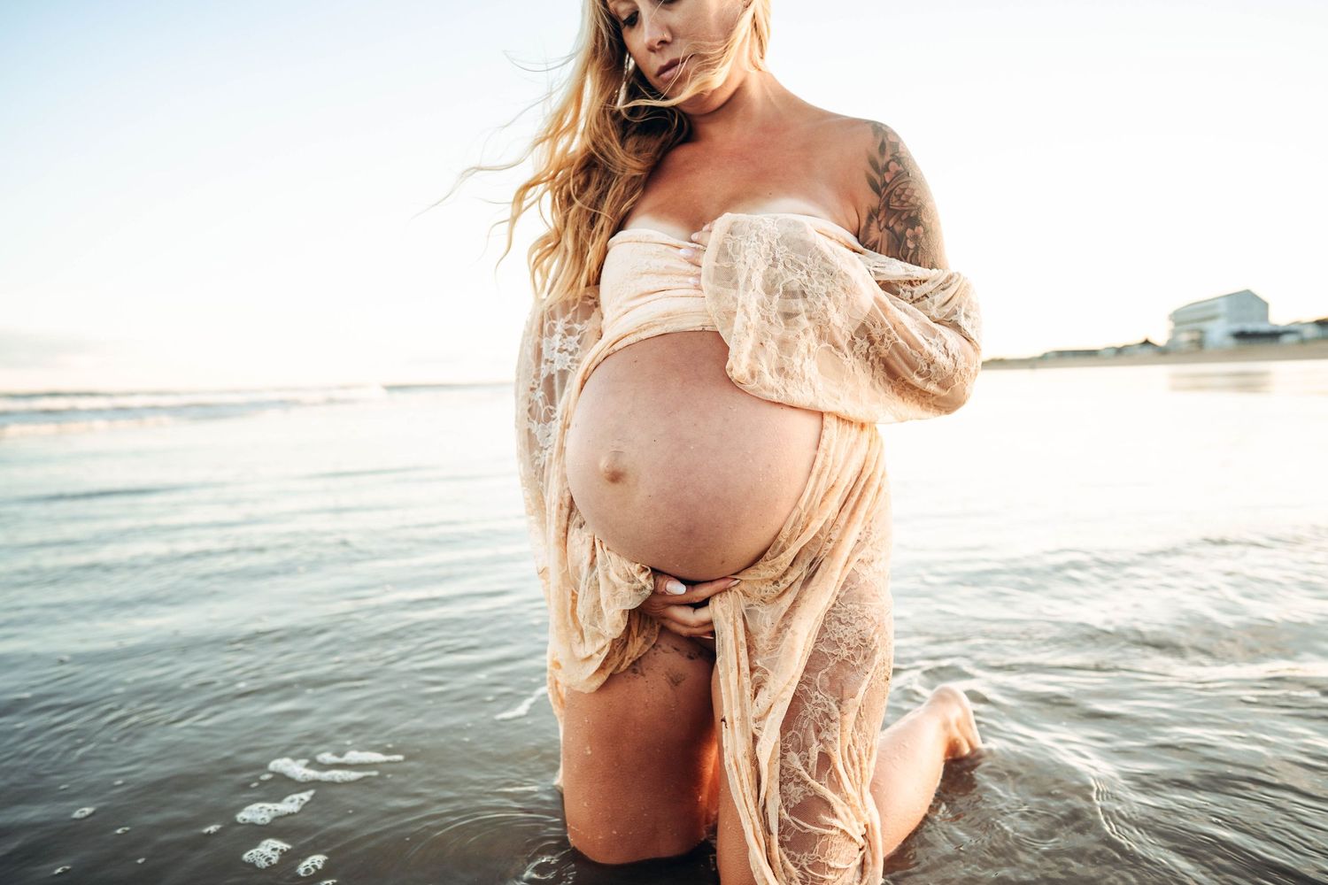 Pregnant woman kneeling in ocean in lace dress.