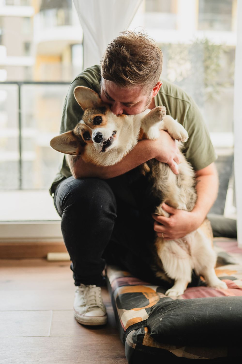 Person in green shirt hugging and cuddling with a happy Corgi dog in a bright room.