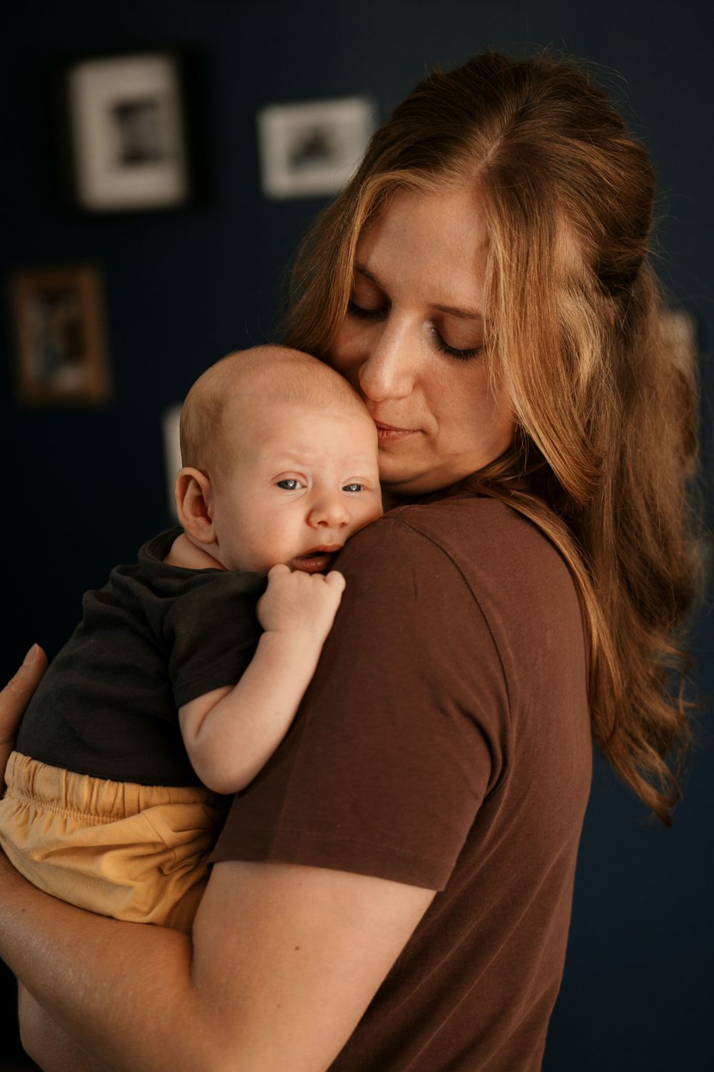 Tender moment between parent and baby against dark wall with framed photos.