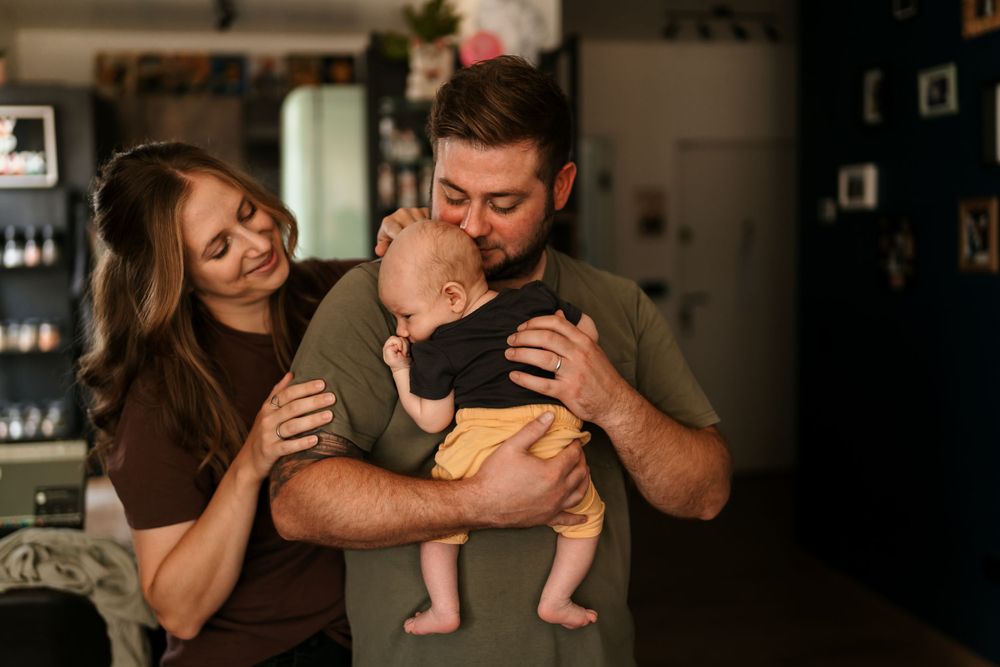 A couple cuddles with their infant baby in a dimly lit room with dark walls.
