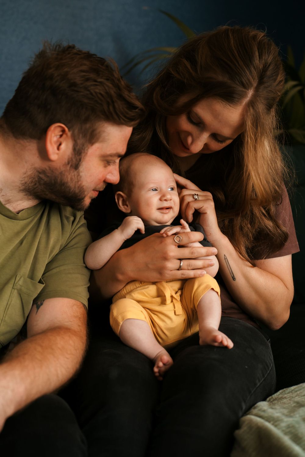 A family shares tender moment with baby wearing yellow outfit on dark couch.