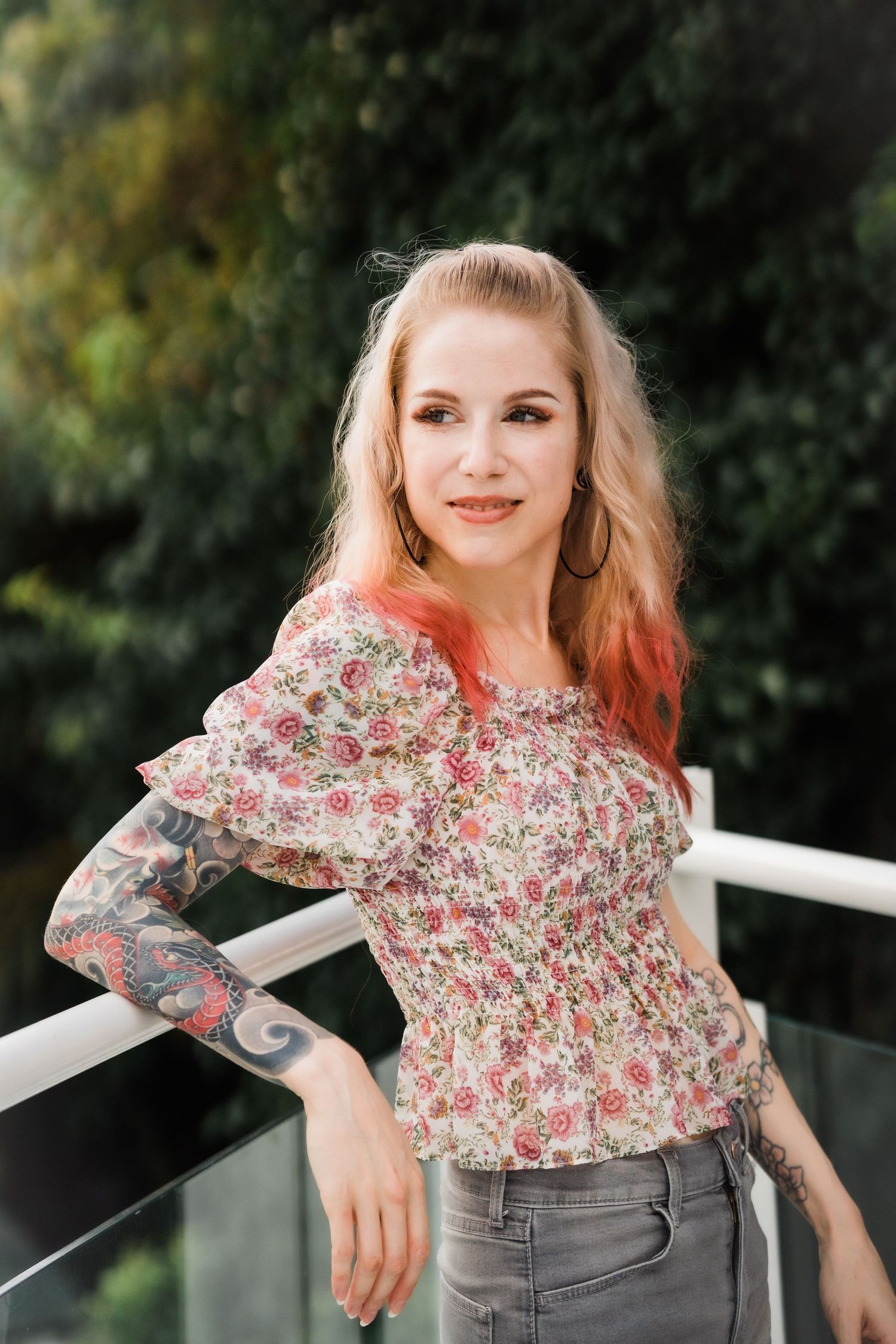 Woman in floral blouse with tattoo poses against outdoor railing.