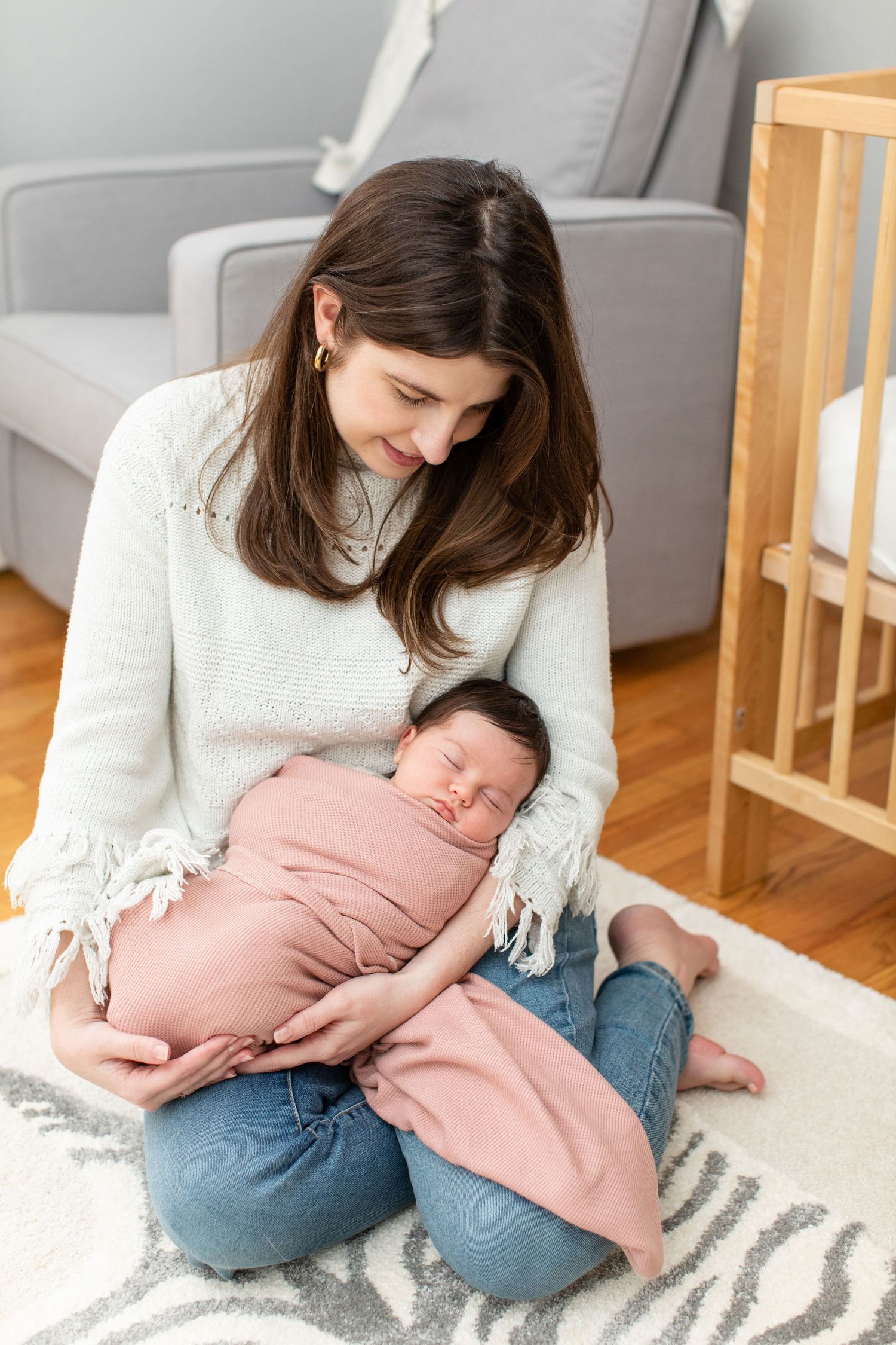 Woman in white sweater sitting cross-legged on rug holding sleeping baby in pink wrap.