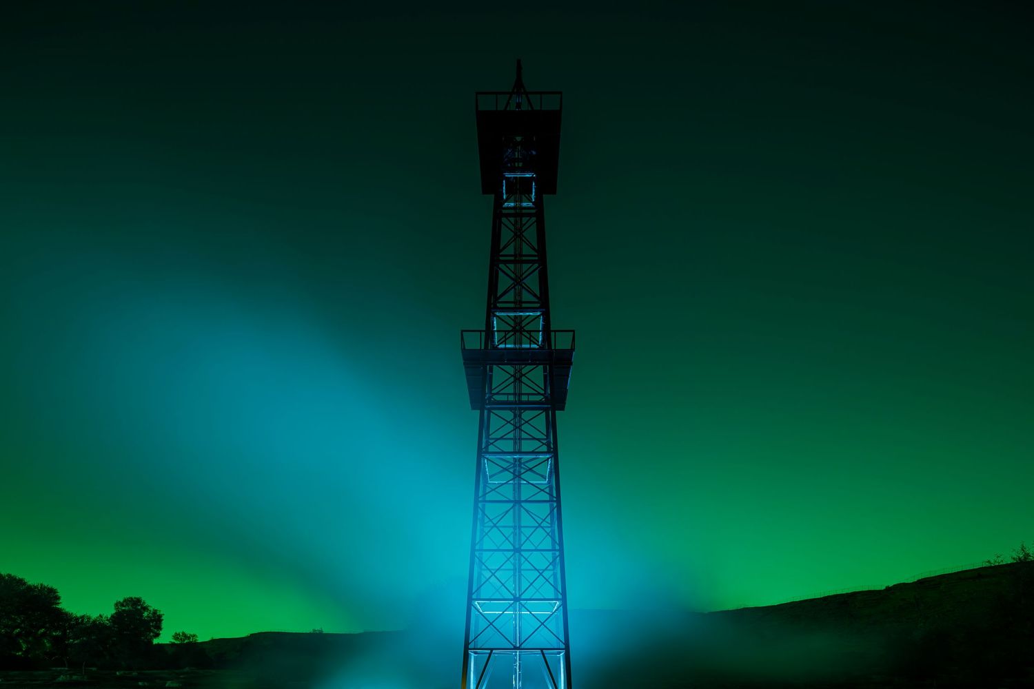 Communication tower silhouetted against dramatic turquoise and green night sky.