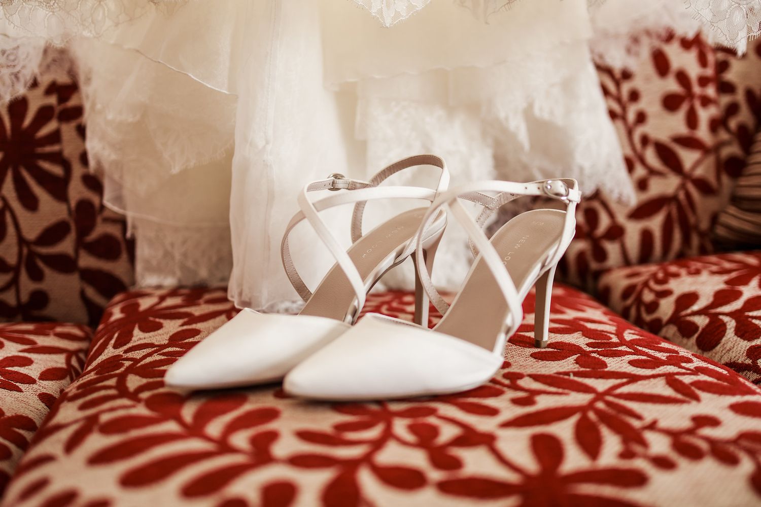 White wedding shoes with lace details rest on a decorative red and white patterned surface.