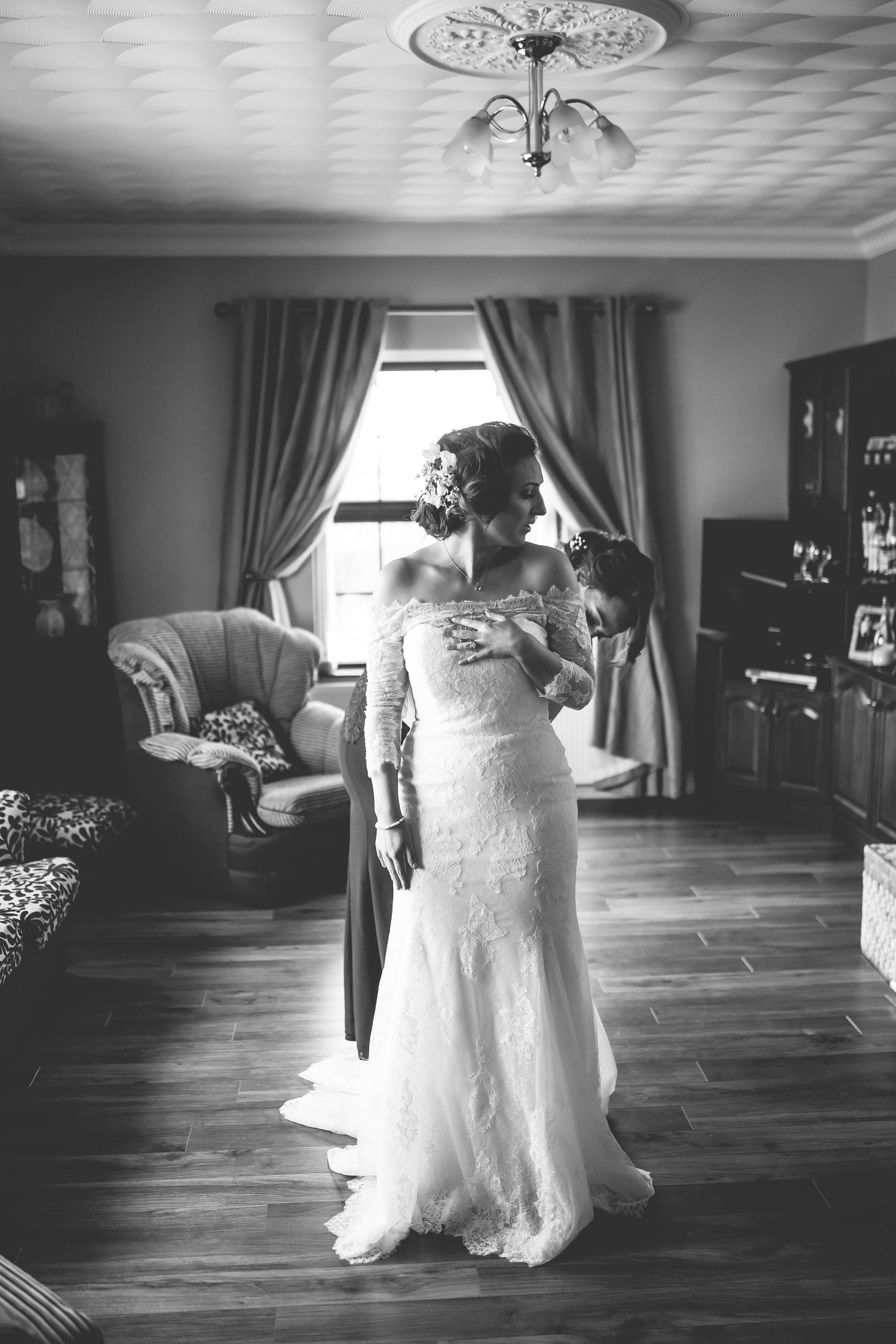 Black and white photo of wedding dress preparations in a sunlit room with hardwood floors.