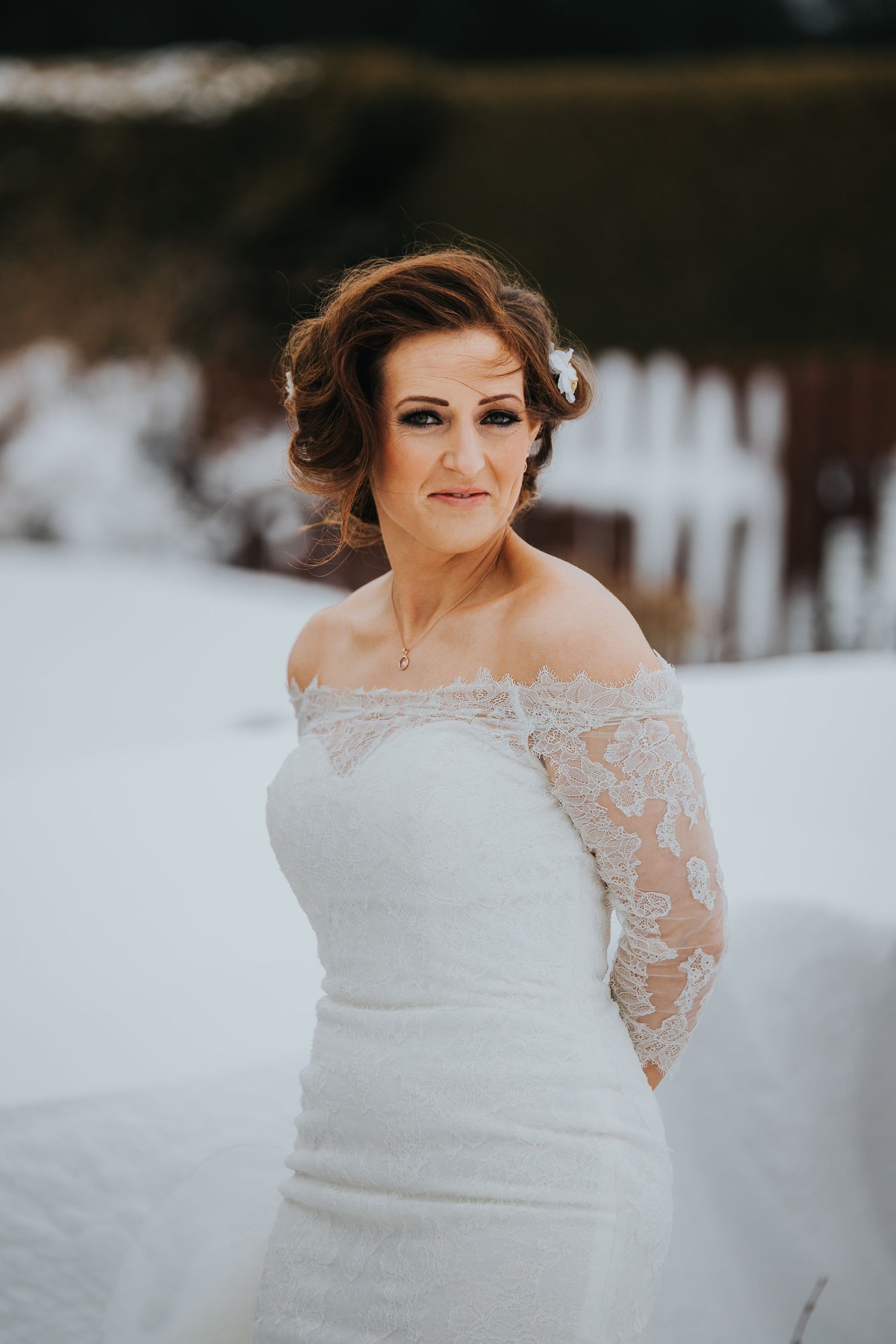Bride in lace-trimmed wedding dress poses against snowy winter backdrop.