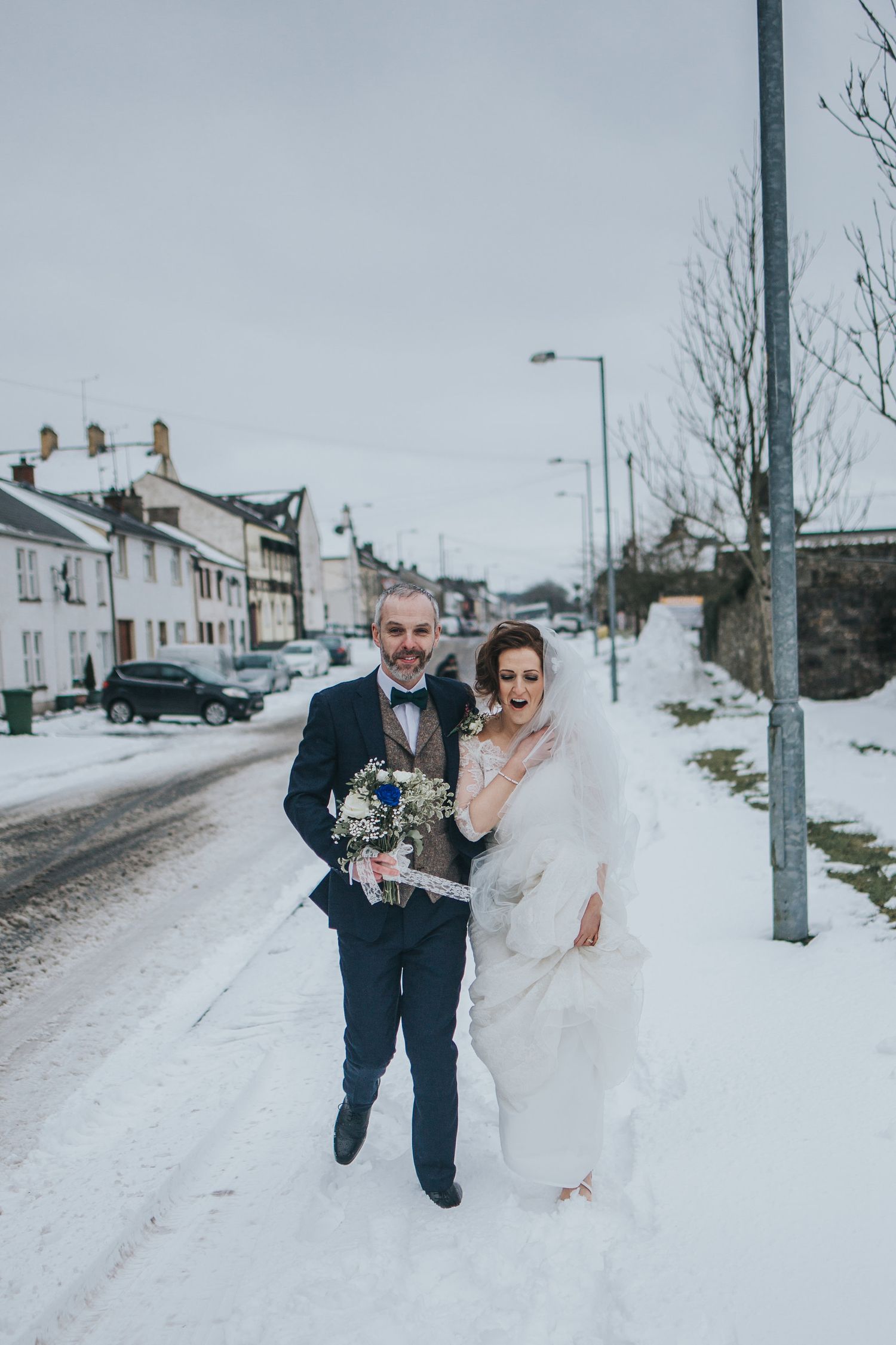 Newlyweds run playfully through snow-covered street during winter wedding photoshoot.