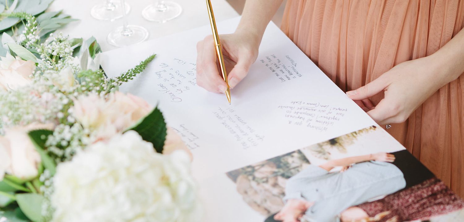 Person in a peach dress writes in a wedding guest book beside a floral centerpiece.