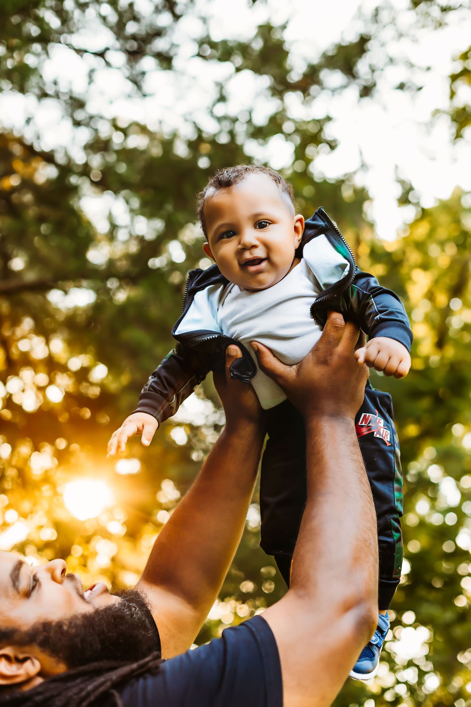 Baby in dark jacket is lifted playfully against golden sunset background.