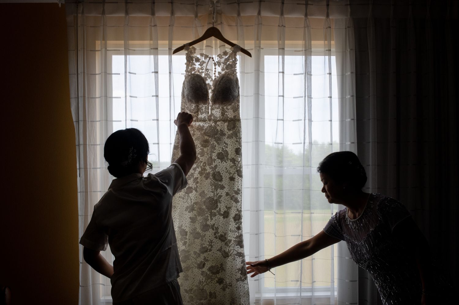 Silhouettes admiring wedding dress hanging in window light.