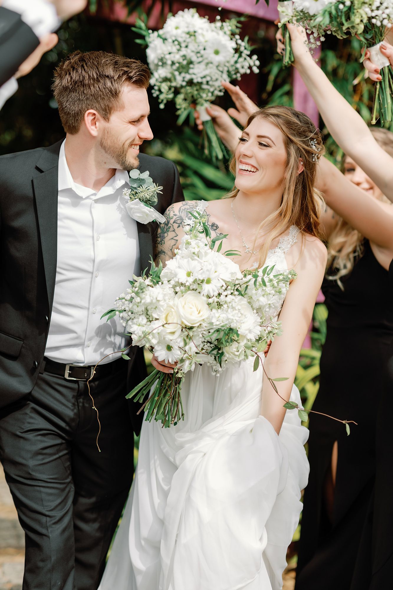 A joyful wedding couple laughs together while holding white floral bouquets among celebrating guests.